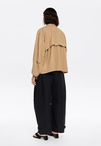 Lichtbeige oversized jack met gelaagd achterontwerp, gecombineerd met zwarte wijde broek en sandalen met hak, die een relaxte pasvorm tonen.