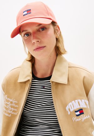 Korallfarget caps med "TOMMY JEANS"-logo, kombinert med en brun semsket jakke. Under er det en svart-hvitrandete skjorte med en liten logo.