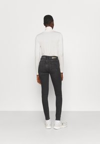 Jeans ajustados negros de cintura alta con un logo dorado en la parte posterior de la cinturilla, combinados con un suéter de cuello alto blanco ajustado y zapatillas blancas.