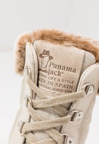 Beige semsket støvel med en myk, tan-farget pelsforing. Snøring med metalløyer og et merket patch som leser "Panama Jack, laget i Spania."