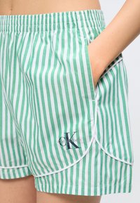 Άτομο που φορά κοντό παντελόνι Calvin Klein με κάθετες πράσινες και λευκές ρίγες και ελαστική μέση, με το χέρι στην πλαϊνή τσέπη, κοντινή άποψη.