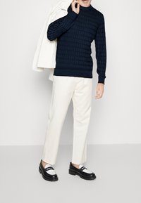 Marineblauwe getextureerde gebreide trui, crème broek, zwart-witte loafers. Model houdt een wit jasje vast, staand tegen een lichtgrijze achtergrond.