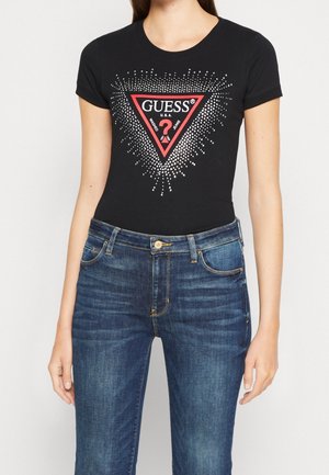 Kvinde iført en sort Guess T-shirt med et rødt trekantslogo og blå jeans mod en ensfarvet hvid baggrund.