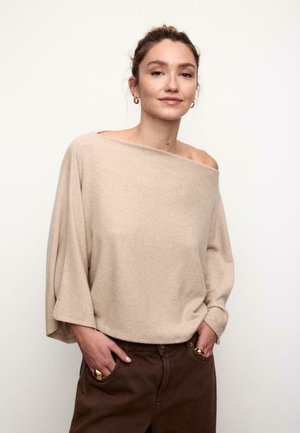 Beiger Strickpullover mit Carmenausschnitt und weiten Ärmeln, weicher Textur und lockerem Sitz, kombiniert mit dunkelbraunen Hosen. Akzente in goldfarbener Hardware.