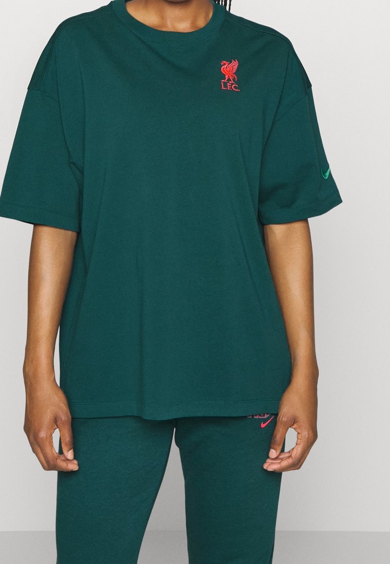 Personne portant un maillot vert foncé du Liverpool FC et un pantalon assorti avec le logo rouge de Liverpool sur la poitrine et le swoosh rose de Nike sur le pantalon.