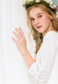 Vestido blanco fluido con mangas abullonadas, cabello rubio ondulado y una corona de flores. Lleva una pulsera plateada en la muñeca. El fondo presenta cortinas blancas suaves.