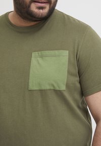 Oliven grøn bomulds t-shirt med en kontrastfarvet lysegrøn brystlomme. Blød tekstur, rund hals, korte ærmer og pæne syninger.