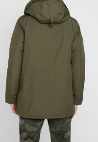 Chaqueta acolchada verde oliva con capucha, que presenta una textura suave y un corte recto. Incluye una cinta en la parte trasera y detalles de diseño minimalistas.