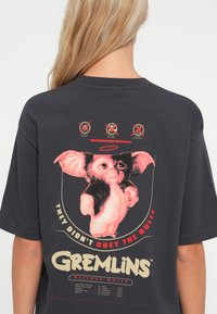 Pelēks pārsistedzēts T-krekls ar krāsainu Gremlins grafiku. Teksts saka: "Viņi neievēroja noteikumus" ar maziem ikoniem augšā.