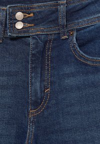 Temno modri jeans z visokim pasom, ki imajo dve kovinski gumb in oranžno šivanje ter sprednji žep. Gladka tekstura in klasičen dizajn.