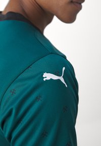 Teal sportströja med texturerad tyg, svart krage och vit Puma-logotyp på axeln. Litet bourgogne-mönster synligt överallt.