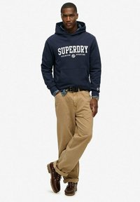 Marineblauwe hoodie met "SUPERDRY" tekst, beige chinos en bruine veterschoenen. Gelaagd denim overhemd zichtbaar bij de kraag.
