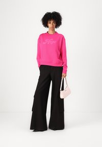 Felpa rosa brillante con logo "Hugo", abbinata a pantaloni neri a gamba larga. Il modello tiene una borsa rosa chiaro con una tracolla sottile.