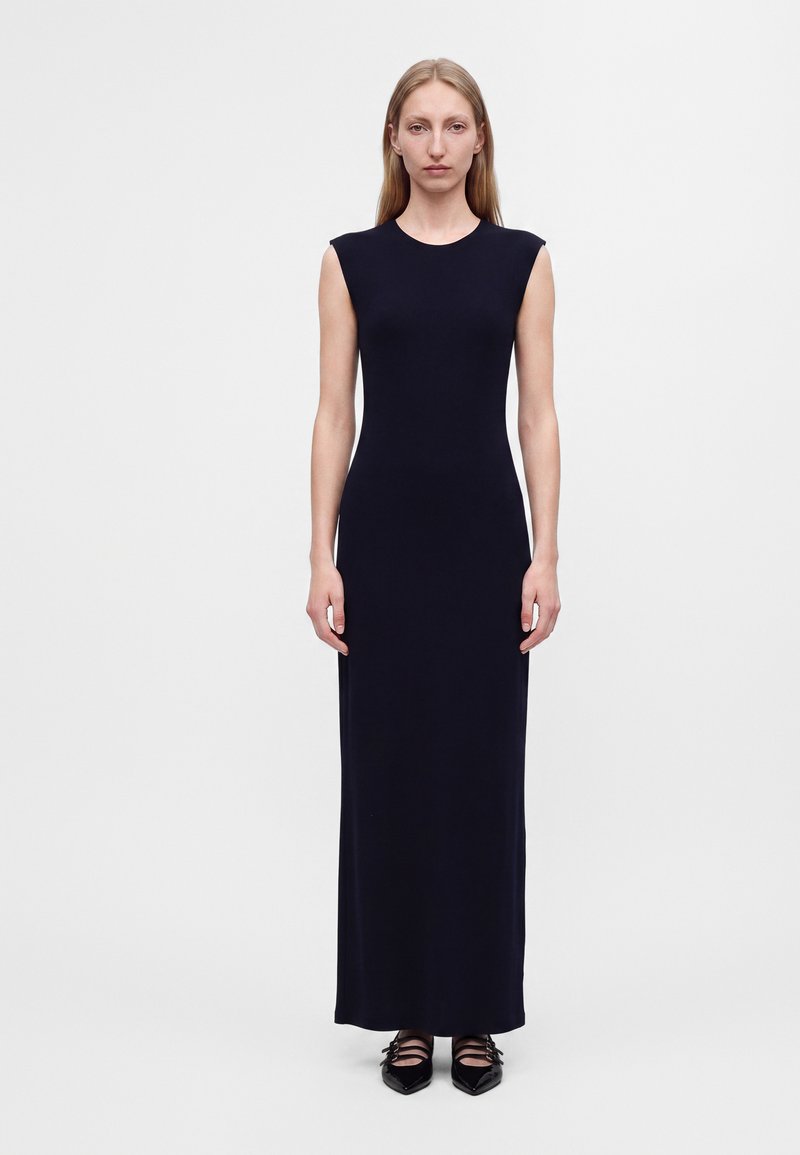 Femme aux cheveux blonds raides portant une robe maxi noire sans manches et des chaussures noires à bouts pointus, debout devant un fond clair uni.