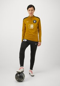 Yellow long-sleeve sports top with black accents, logo on chest; black athletic pants, white cleats with red accents, player standing on a black ball.