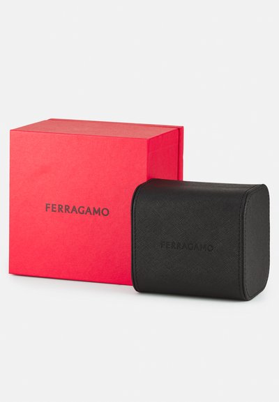 Black leather round case with textured finish, embossed logo, beside a red cardboard box with printed black logo.