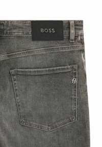 Szare dżinsy z denimu mają teksturowaną czarną skórzaną łatkę z nazwą marki "BOSS". Tylna kieszeń ozdobiona jest metalowym detalem.