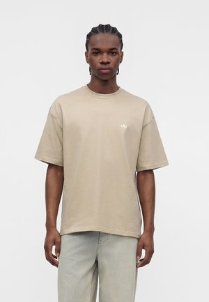 Ung mand med fletninger iført en beige Adidas t-shirt og lyse jeans, stående mod en ensfarvet hvid baggrund.