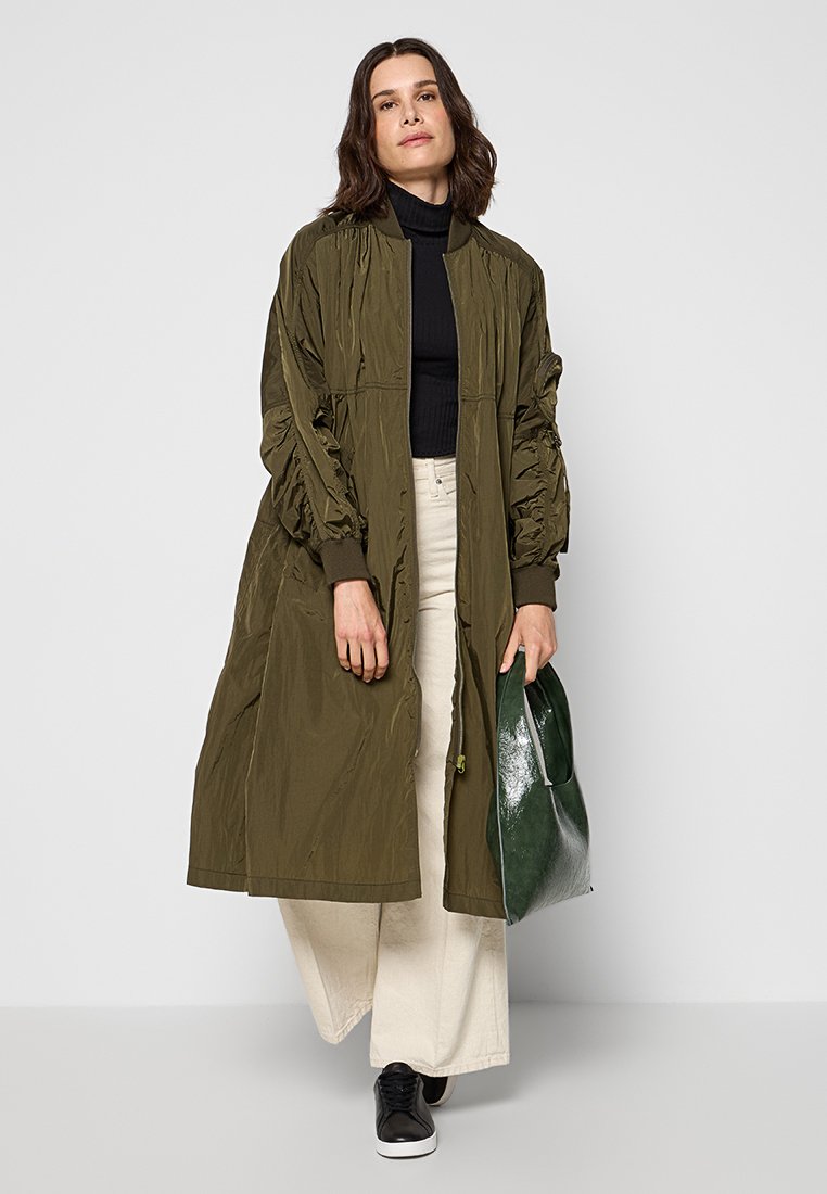 Oliwkowy długi bomber z marszczonymi rękawami, zestawiony z czarną bluzką i jasnobeżowymi spodniami o szerokich nogawkach, trzymający błyszczącą zieloną torebkę.