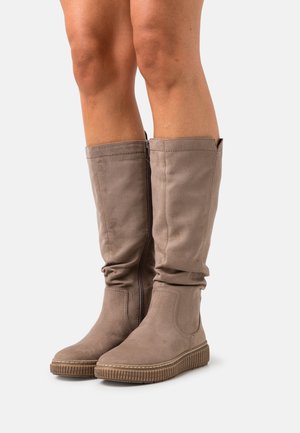 Polven korkeuteen ulottuvat taupenväriset mokkanahkasaappaat, joissa on sivuvetoketju, tasainen uurrettu pohja, käytetty paljaiden jalkojen kanssa yksinkertaista vaaleaa taustaa vasten.