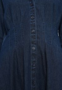 Denimskjorte i mørkeblå, med knapper foran med syv knapper, lodrette syninger og diskrete plisseringsdetaljer.