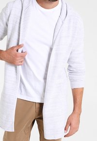 Lys grå, struktureret cardigan med hætte, lange ærmer og åben front. Båret over en hvid t-shirt og kombineret med beige bukser.