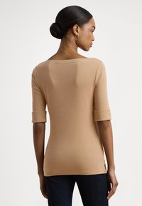 Μπεζ, εφαρμοστό τοπ κατασκευασμένο από μαλακό ύφασμα, με φαρδιά λαιμόκοψη, μανίκια τριών τετάρτων με ρολομάνικες και πλαϊνές σχισμές στο τελείωμα.