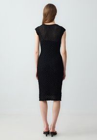 Robe noire en tricot avec un motif texturé, des manches courtes et une silhouette ajustée. Longueur mi-mollet avec un détail en dentelle au dos.