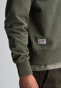 Close-up van een persoon in een olijfgroen sweatshirt met een geribbelde manchette en een klein label "Butcher's Clothing Company" nabij de taille.