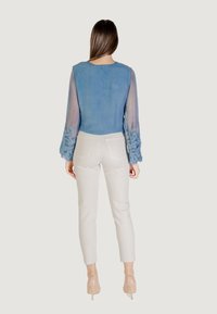 Blusa de manga larga con un frente azul, similar a ante, y mangas transparentes, decorada con intrincados bordados azules en los puños. Combinada con pantalones claros.
