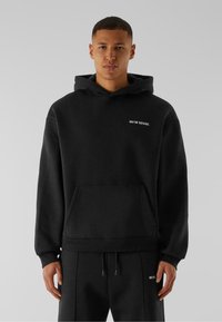 Sweatshirt à capuche noire en tissu doux, avec une poche avant et un texte de logo blanc sur la poitrine gauche. Présente des poignets et un ourlet côtelés.