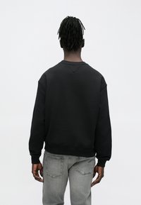 Persona con rastas que lleva una sudadera negra y vaqueros grises desgastados, de pie de espaldas contra un fondo claro y sencillo.