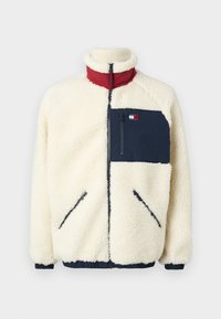 Giacca in pile color crema con colletto rosso, tasca blu navy e zip. Caratterizzata da polsini e orlo elastici, con un logo sulla tasca del petto.