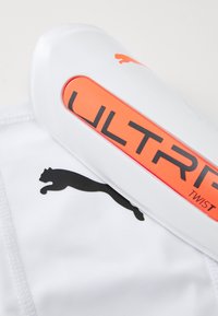Bílé sportovní chrániče holení s oranžovým a černým logem s nápisem "ULTRA TWIST". Obsahuje černé logo Puma a hladký povrch.