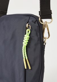 Sac en tissu bleu marine avec une surface texturée, doté d'une fermeture éclair dorée, d'un tirant de fermeture éclair en paracorde vert lime et d'une sangle noire réglable.
