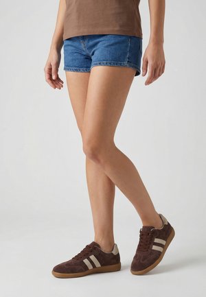 Persoon die bruine sneakers met beige strepen draagt, blauwe spijkershorts en een bruin topje, staand op een lichtgrijze achtergrond.