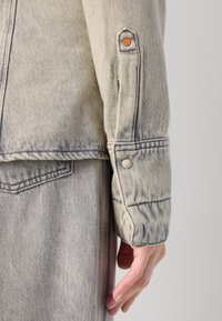 Manica di giacca in denim grigio chiaro con chiusura a bottone e un'applicazione decorativa sul polsino; texture liscia con una leggera sfumatura.