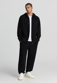 Felpa nera con cappuccio e tasche frontali, abbinata a pantaloni sportivi neri, presentata con scarpe da ginnastica bianche. Design minimalista, tessuto misto cotone.