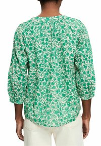 Femme portant un chemisier à manches longues vert et blanc à motif floral, avec des manches bouffantes trois-quarts, vue de dos.