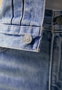 Salopette in denim azzurro con dettagli cuciti, pince sulle spalle e un bottone in metallo argentato con il testo del marchio.