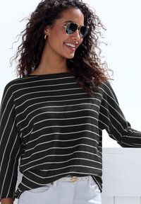 Lächelnde Frau mit lockigem Haar, die eine aviator Sonnenbrille, einen schwarz-weißen gestreiften Pullover, weiße Hose und einen weißen Gürtel mit einer goldenen Schnalle trägt.