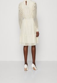 Kramfärgad klänning med strukturerad tyg, spetsdetaljer, långa ärmar och en utsvängd kjol, tillsammans med vita högklackade skor.