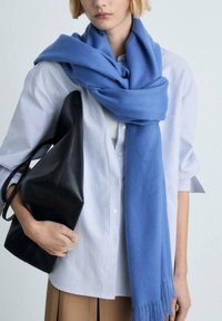Light blue scarf made of soft fabric, draped around the neck. Striped button-up shirt underneath and a black leather bag held in one hand.