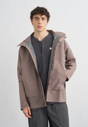 Hombre joven con una chaqueta con capucha color topo sobre una camiseta henley gris oscuro y pantalones cargo oscuros, de pie frente a un fondo claro y sencillo.