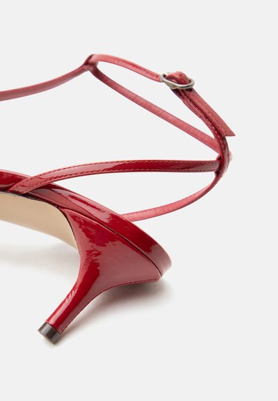 Κόκκινο σανδάλι με τακούνι κιττέν από λουστρίνι, με λεπτό λουράκι στον αστράγαλο και μικρή ασημένια αγκράφα σε λευκό φόντο.