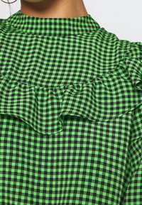 Green and black checkered top featuring a ruffled neckline. The fabric displays a grid pattern with texture from the ruffle detail.