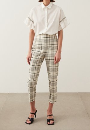 Valkoinen nappimallinen paita, jossa on liehukahihat, yhdistetty beigeihin ruudullisiin korkeavyötäröisiin housuihin ja mustiin nauhalla varustettuihin korkokenkiin.