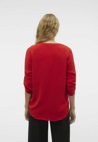 Maglietta rossa a maniche lunghe con un motivo testurizzato, scollo rotondo e orlo arrotondato, abbinata a pantaloni neri, vista da dietro.