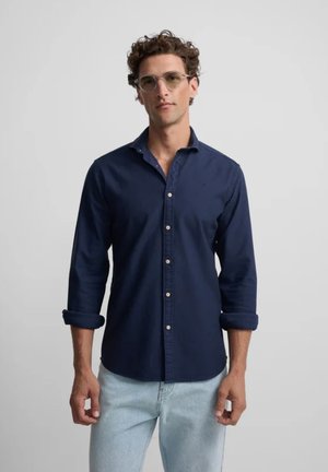 Hombre con cabello rizado y gafas, vistiendo una camisa azul oscuro abotonada con las mangas remangadas y jeans azul claro, sobre un fondo liso.