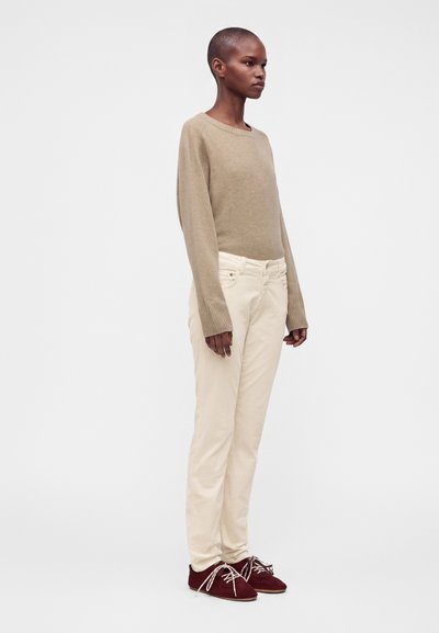 Personne aux cheveux courts portant un pull beige, un pantalon écru et des chaussures à lacets rouge foncé, debout devant un fond clair uni.